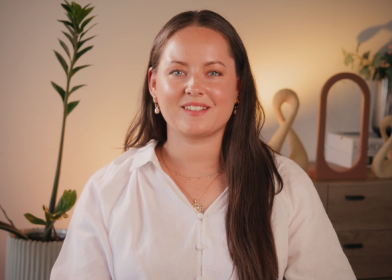 smiling woman in white shirt with long hair, green plant in background, warm lighting creating inviting atmosphere, friendly portrait with elegant decor and soft lighting, showcasing personal style and home setting